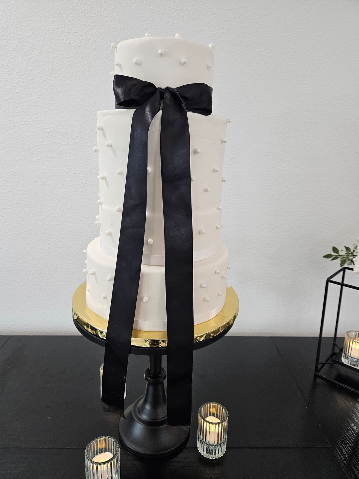 Three tier white wedding cake with sugar pearls and a long black satin bow on a black cake stand