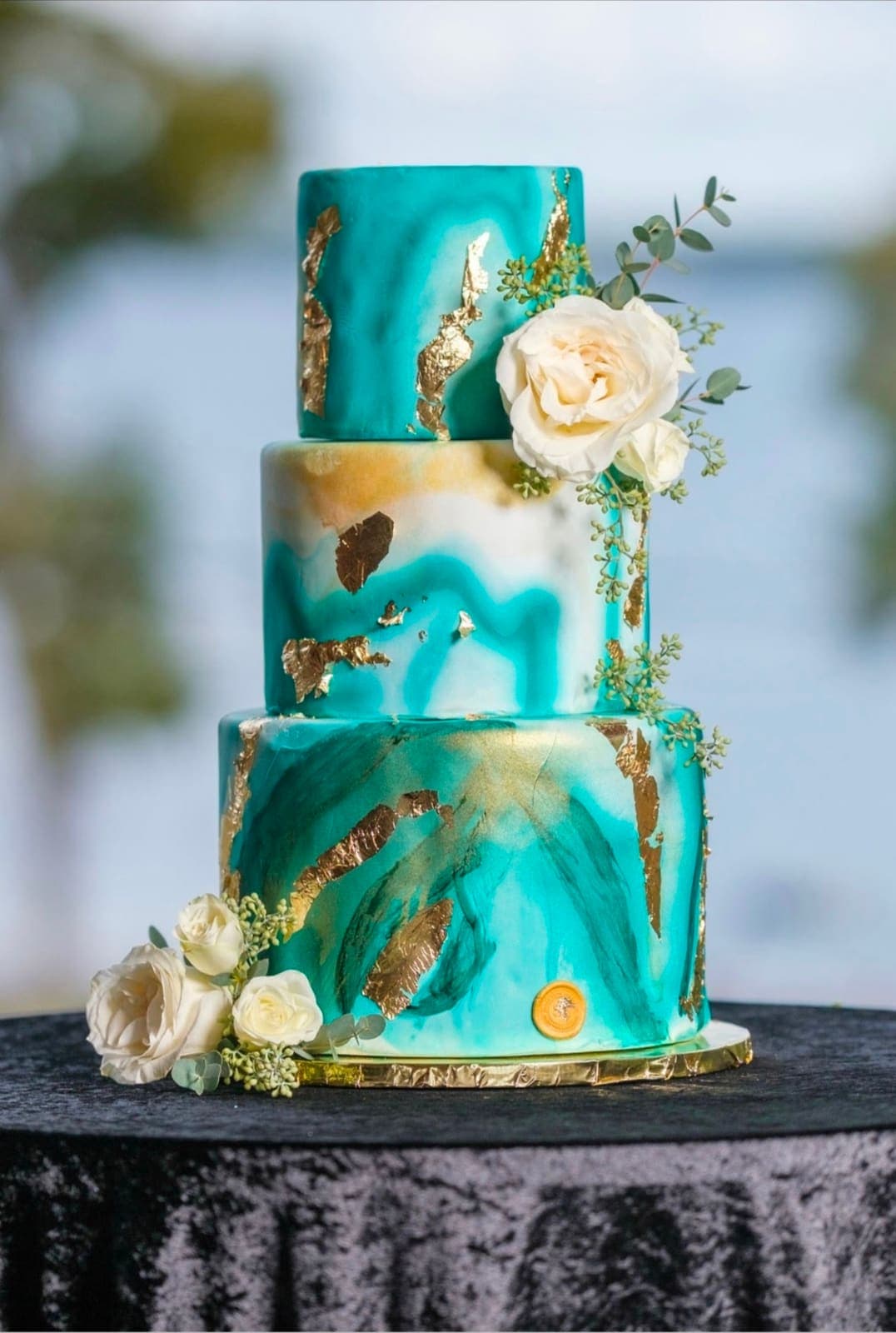 Three tier teal and white marbled wedding cake with gold leaf and white roses, set beside the water