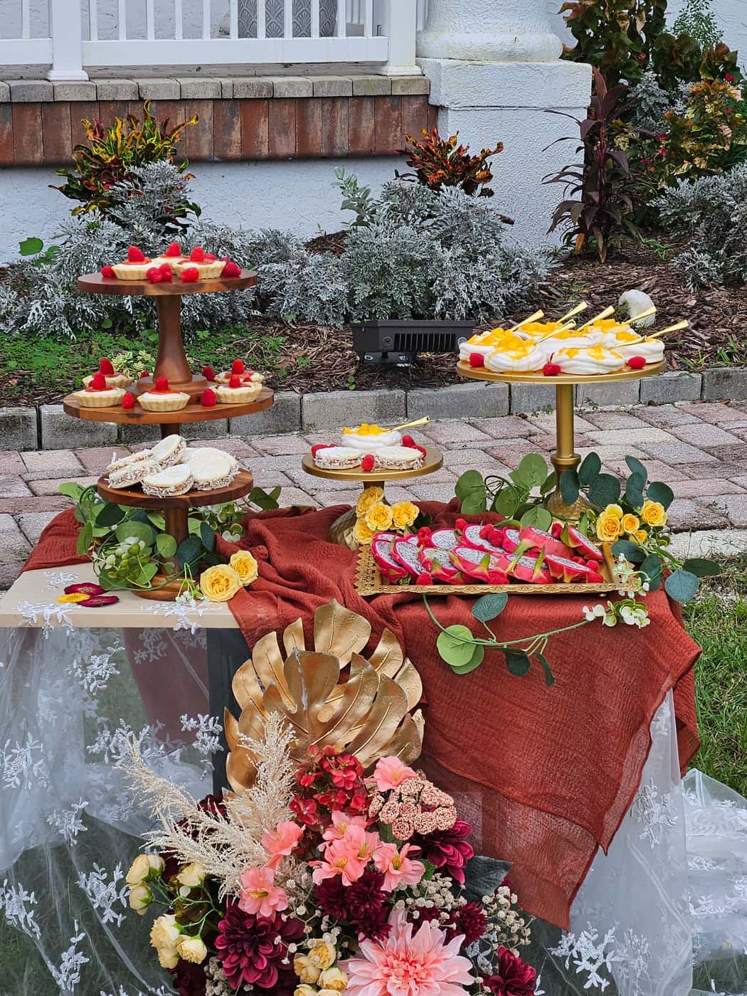 Styled outdoor dessert installation with tartlets, pavlovas and dragon fruit on wood and gold stands, draped in rust fabric with fresh flowers