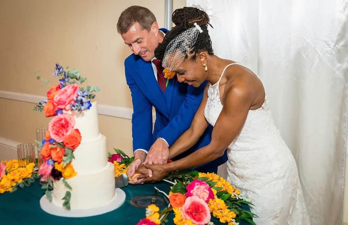 A couple cutting a three tier white wedding cake dressed with orange, coral and blue flowers