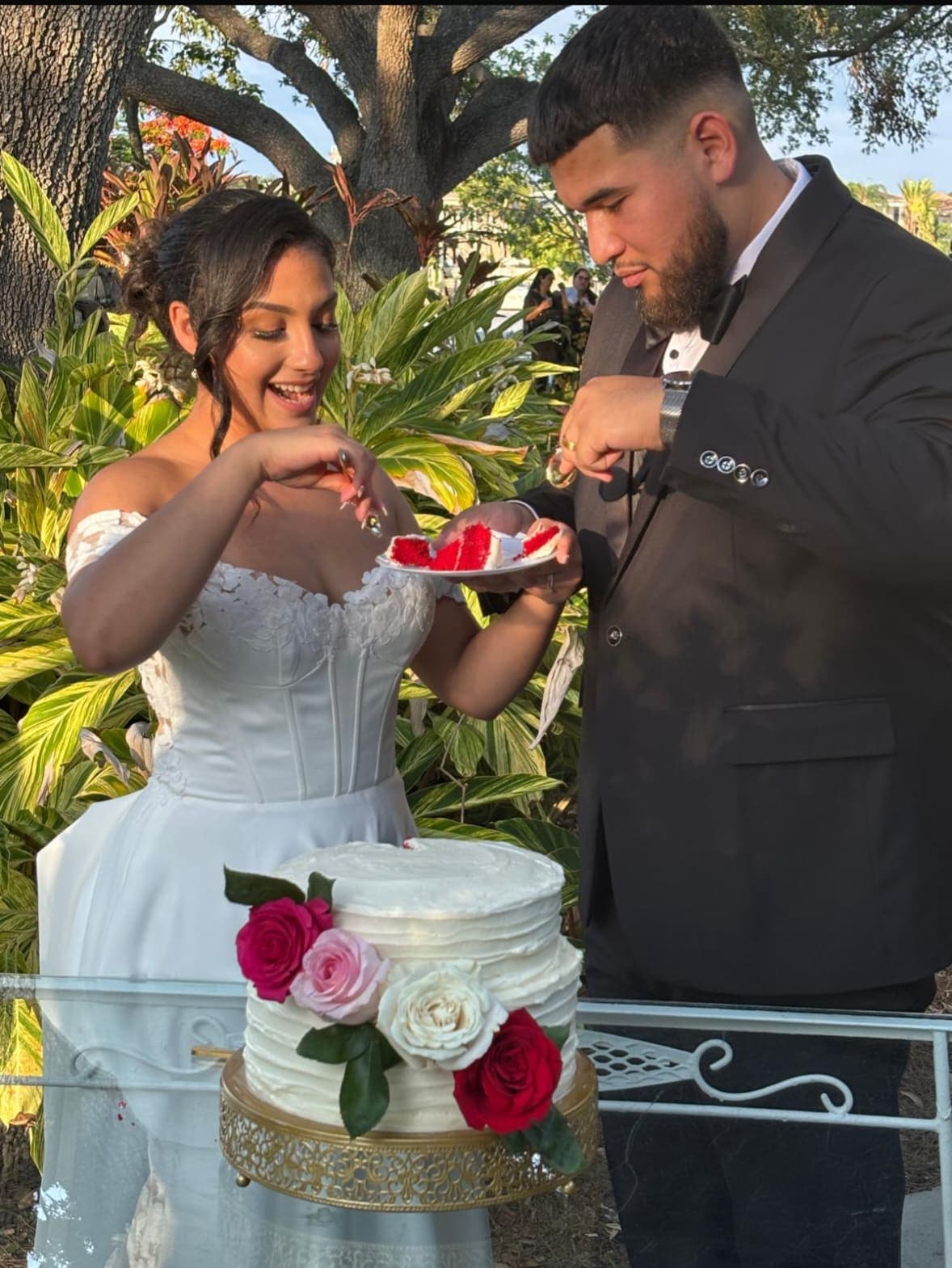 A bride and groom sharing a slice of cake behind a two tier white cake with red and pink roses