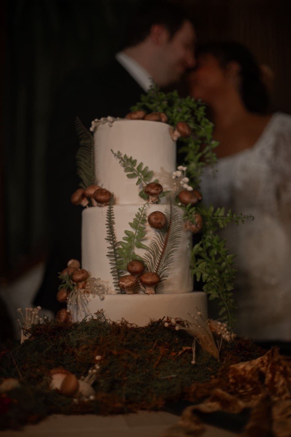 Three tier white wedding cake decorated with ferns and mushrooms on a bed of moss