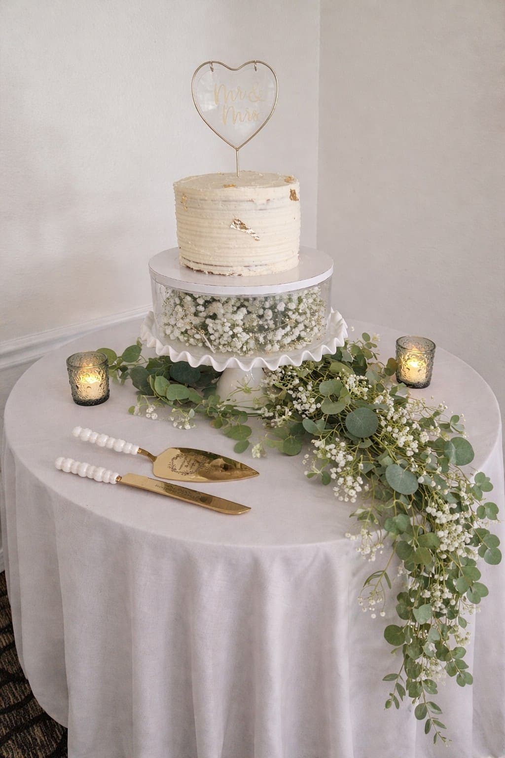 One tier textured buttercream cake with gold leaf on a stand filled with baby's breath, with eucalyptus across the table