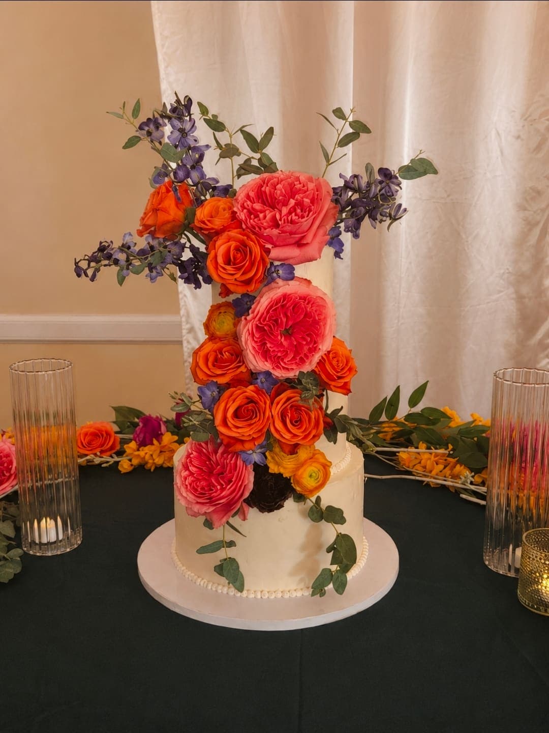 Three tier white wedding cake covered in orange roses, coral garden roses and blue flowers